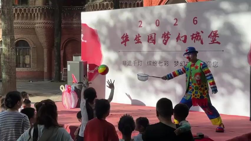 A clown in a colorful costume and blue hat is performing on a stage in front of a crowd of people, including children and adults. He's holding a net and appears to be attempting to catch a small ball. The stage is set up outdoors, with a brick building visible in the background. A large white banner behind the clown has red lettering and appears to be advertising an event called "2026 华生园梦幻城堡." The crowd is watching the performance, with some people raising their hands. An audio speaker sits to the left of the stage. A woman walks behind the clown during the show.