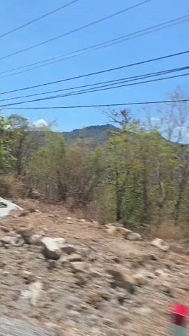 This video captures a bright, sunny daytime scene of a dry, mountainous landscape, likely viewed from a moving vehicle. The terrain is characterized by rocky, dusty ground and hills covered in sparse, dry vegetation. In the foreground, numerous rocks are scattered across the ground, with some small green bushes visible. Power lines stretch across the clear blue sky, and a wooden utility pole briefly appears on the right side of the frame as the camera pans. The background features rolling hills and mountains covered in a mix of green and dry brown foliage, suggesting a warm and possibly arid climate. The bright sunlight and lack of clouds indicate good weather. No people are visible, and there is no discernible text in the video. The scene depicts a rural or semi-rural area, consistent with the provided context of Phan Sơn, Vietnam.