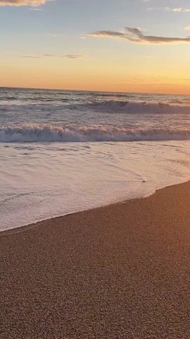 The video shows a beach at sunset. The sun is setting on the horizon, creating a golden reflection on the water and the beach. The sky is filled with orange and pink hues, with some clouds scattered across the sky. The waves are gently rolling in, and the beach is made of small pebbles. There are no people visible in the video.