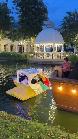 A family of four enjoys a pedal boat ride on a small pond at dusk. The boat is shaped like a yellow duck, with a white roof and colorful accents. Inside the boat, a woman and two children pedal, while another woman stands on a nearby wooden boat, looking over at them. The setting appears to be a park or resort, with a domed building and trees visible in the background, all illuminated by string lights. The overall atmosphere is peaceful and festive.