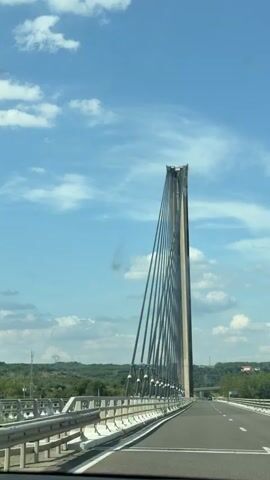 The video shows a view from a vehicle driving across a modern cable-stayed bridge. The primary subject is the bridge's towering concrete pylon, from which numerous steel cables fan out, supporting the bridge deck. The road surface is visible, with white markings and barriers on either side. In the background, a lush green landscape with rolling hills and trees can be seen under a bright blue sky dotted with fluffy white clouds. The time of day appears to be daytime, likely midday or early afternoon, given the clear visibility and sunlight. The weather seems fair and pleasant. The location is identified as La Baume-d'Hostun, France. The overall impression is one of engineering prowess and a scenic drive. No people are visible in the shot. There is no visible text in the media file.