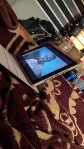 A tablet is propped up on a patterned blanket, displaying a video. The video shows different people on screen. The tablet is on a surface that appears to be a bed, covered with a blanket with a maroon and cream colored pattern. In the background, there are bags and a wall with black and white striped wallpaper. The camera is shaky and blurry at times.
