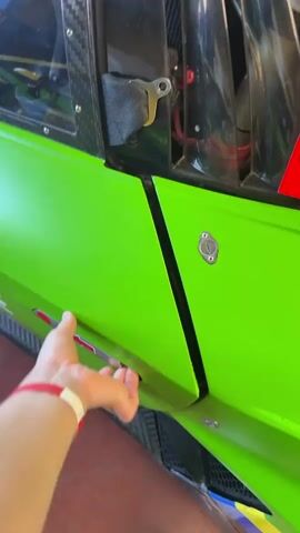 A hand opens the driver's side door of a green race car to reveal the vehicle's interior. The car has a racing-style steering wheel, a series of dashboard switches, and a racing seat with a harness. The door is then closed.