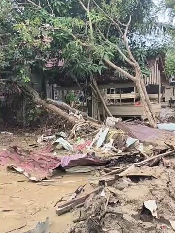The video shows the aftermath of a flood. Mud and debris cover the ground around houses. There are pieces of metal, wood, and other building materials scattered everywhere. A tree lies uprooted next to one house. Laundry hangs on a line outside another house. The scene is one of devastation.