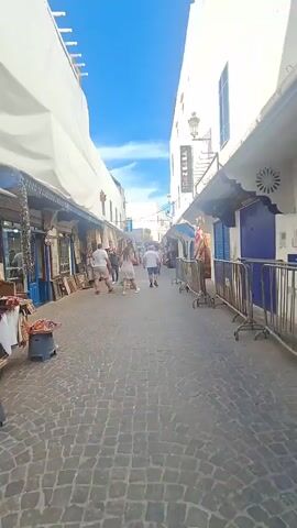 A vertical video shows a narrow cobblestone street lined with shops on both sides. The shops appear to be selling textiles and other goods. Several people are walking down the street, mostly away from the camera. The buildings are whitewashed, and the sky is blue with a few scattered clouds. A sign on the right-hand building reads "MENZEL." The overall impression is of a busy market street in a Mediterranean or North African town.