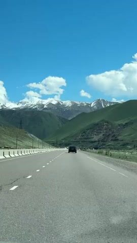 A daytime video captures a black sedan driving on a paved highway through a majestic mountainous landscape in Cholok, Kyrgyzstan. The road features two lanes in the direction of travel, separated by dashed white lines, and is bordered by concrete barriers. On either side, steep green hills and valleys rise, leading to towering snow-capped mountains in the background. The sky is a vibrant blue with scattered white, fluffy clouds. Other vehicles, including a white car and a white truck, are visible in the distance. The black sedan in front has a Kyrgyz license plate displaying "03 KG 061 ACI." The scene conveys a sense of serene travel through stunning natural scenery under clear weather conditions.