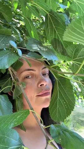 A close-up video shows a woman's face peeking through lush green leaves of a tree. Her dark hair frames her face, and she is looking towards the camera with a neutral expression. The background is blurred, but it appears to be an outdoor setting with water visible. The lighting suggests it is daytime, and the overall mood is natural and serene.