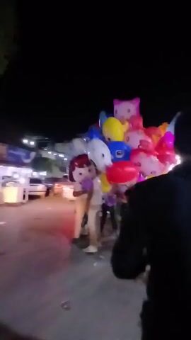 At night, a group of people are walking down a paved road, illuminated by streetlights. A man holds a large bunch of character-themed balloons, including Hello Kitty and other cartoon figures. Other people in the group are dressed in light-colored clothing. In the background, there are parked cars and buildings with lights on. The camera follows the group as they move along the road.