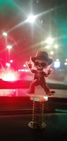 A small, bobblehead skeleton figurine wearing a fedora and a jacket is mounted on a spring and placed on a dashboard inside a vehicle. The figurine is positioned in the center of the frame and is depicted with its hands raised in a rock and roll gesture. The background shows a blurred street scene at night, with red and green lights from traffic signals and streetlights. The lighting suggests that the vehicle is either stopped at an intersection or moving through an area with traffic lights. The overall impression is a playful and slightly edgy decorative item in a car at night.