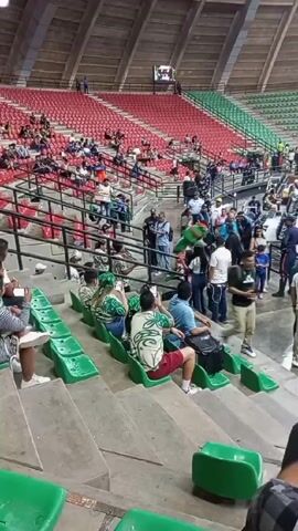 The scene unfolds inside a large indoor stadium with tiers of red and green seats sparsely populated with people. In the lower rows, a group of spectators wearing predominantly green clothing are seated, many holding up their phones to record the event. There is a small crowd of people standing and milling about, including a mascot in a green and white suit. The stadium's ceiling is ribbed and textured, and the overall lighting is somewhat dim. Some spectators are seen moving around, taking pictures, or just sitting and observing the activity.