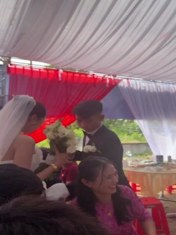 The video captures an outdoor celebratory event, likely a wedding reception, taking place under a large tent adorned with draped white, red, and blue fabric, strung lights, and decorative white paper flowers. The setting is Tay Son Commune, Vietnam, during daylight hours.

Many guests are present, some seated at round tables, while others move through the gathering. A man in a black suit with a bow tie, possibly the groom, is seen holding a bouquet of white flowers, and a woman in a white dress with a veil (likely the bride) is partially visible next to him. Other attendees include women in a pink patterned top, a white top, and a traditional red Vietnamese áo dài.

At the front of the tent, a stage area is set up with a sound system, large speakers, and musical instruments including a keyboard. Musicians are performing, and a person is singing in front of a large screen displaying colorful graphics. Text on the screen includes "Nguyet Tuan" and "082 099." The background outside the tent shows some greenery and a rural landscape. The atmosphere appears festive and joyful, with guests engaged in the celebration.