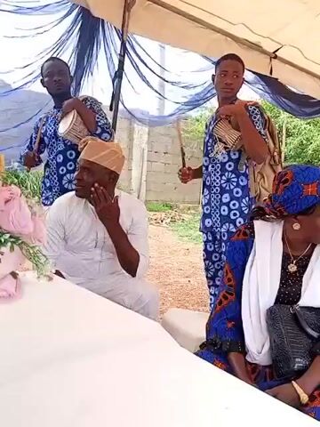 Two men in matching blue and white patterned attire stand in the background under a draped white and blue tent. The man on the left actively plays a talking drum with two sticks. The man on the right holds a talking drum on his shoulder and a single stick, looking forward. In the foreground, a senior man in a white traditional garment and a beige cap sits at a white table on the left, resting his chin on his hand. A senior woman in a blue patterned dress and a white headscarf sits at the table on the right, holding a black purse and looking towards the left. A pink floral arrangement sits on the table to the left. An unfinished brick wall and green foliage are visible in the background.
