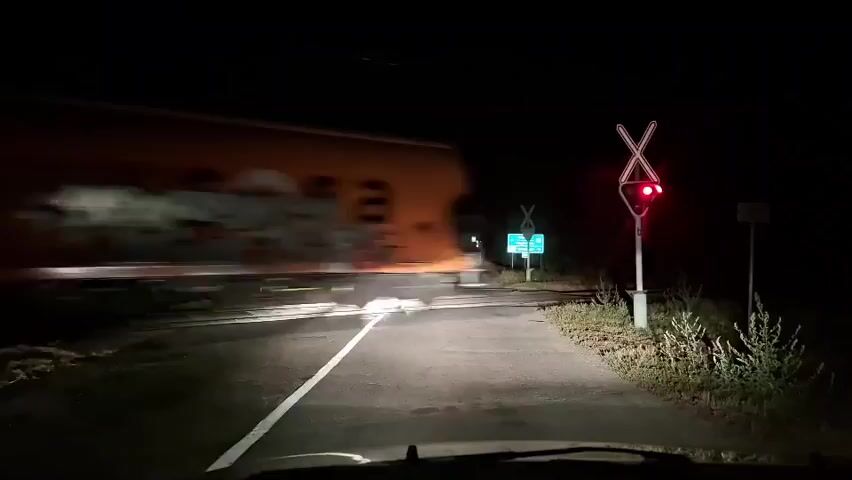 The video is shot from the perspective of someone driving a car at night. The car is approaching a railroad crossing. The crossing signal is lit red, indicating a train is approaching or present. There are other cars ahead with their headlights on. A gate at the crossing appears to be partially down. The road is paved and has white lines. Vegetation is visible on the sides of the road.