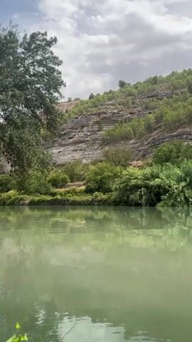 A video shows a serene river with a lush green hillside in the background. The river is calm, with a slight ripple on its surface, reflecting the trees and sky. The hillside is covered in various shades of green foliage, with rocky outcrops visible. The sky is partly cloudy, with patches of blue peeking through. The overall impression is one of natural beauty and tranquility.
