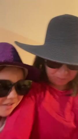 Two individuals, one wearing a purple hat and sunglasses and the other a black hat and sunglasses, are close together. Both are wearing bright pink shirts. The video is shaky and blurry, giving the impression of quick movement or a close-up, handheld shot. The background appears to be a plain, light-colored wall, possibly in a room. The individuals seem to be interacting with each other, possibly taking a selfie or recording a short video.