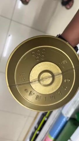 The video begins with a close-up of a gold-colored dumbbell. The camera then pans out to show a person's arm holding the dumbbell. The person is wearing a black watch on their left wrist. In the background, two people are visible standing on a tiled floor, wearing sandals. The environment appears to be an indoor space, possibly a store. The video is handheld and the lighting is consistent throughout.