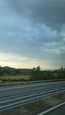 This video shows a roadside view from a moving vehicle on a highway under a cloudy sky. The highway has multiple lanes separated by a barrier and white lines. On the right side of the highway, there is a grassy shoulder with trees and distant hills. Several vehicles are visible on the highway, including a large truck with a blue trailer and a white van. The sky is overcast with dark clouds, suggesting potential rain or an approaching storm. The overall impression is of a journey through a rural landscape with impending weather.