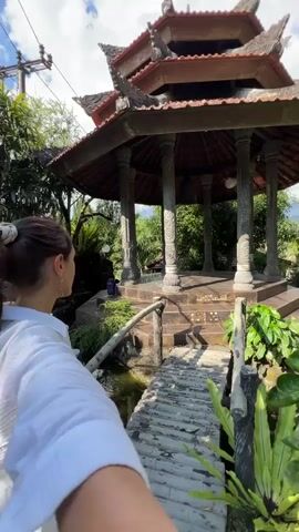 The video shows a woman from the chest up in the foreground, wearing a white top. She is looking upwards and to her right, with her hair pulled back in a ponytail. In the background, there is a gazebo with ornate pillars and a red tiled roof. A bell hangs in the center of the gazebo. The gazebo is surrounded by lush green foliage and trees. A wooden bridge crosses a body of water in front of the gazebo. The scene appears to be outdoors during the daytime, with sunlight filtering through the trees. The camera is held at a low angle, looking up towards the gazebo and the woman.