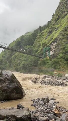 A wide, muddy, fast-flowing river dominates the foreground of the video. The river is filled with debris, including rocks and branches, indicating a recent flood or heavy rainfall. On the left bank of the river, a large, dark boulder is partially submerged in the water. The riverbed is rocky and uneven. In the mid-ground, a suspension bridge stretches across the river, connecting the two banks. Several people can be seen walking across the bridge. On the right bank of the river, a steep, green, and lush mountainside rises sharply. The mountainside is covered in dense vegetation, and the terrain appears rugged and uneven. A small, white structure with a green frame is built into the side of the mountain, near the bridge's landing point. The sky is overcast, with a light mist or fog obscuring the mountain peaks in the distance. The overall atmosphere of the video suggests a wild and potentially dangerous natural environment, possibly experiencing a period of heavy rain or flooding.