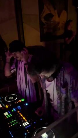 The video shows two DJs working at a nightclub. The DJ on the left wears a black beanie and a vertical striped shirt, while the DJ on the right has curly hair, wears headphones around their neck, and sports a patterned shirt over a white tee. They both focus on the electronic DJ equipment in front of them, which has multiple glowing buttons and turntables. Other people are visible in the background, moving slightly, and the lighting is dim, with a purple hue dominating the scene. A poster hangs on the wall behind the DJs.