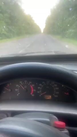 This media file provides a first-person perspective from inside a moving vehicle. The scene is a road with trees lining both sides, suggesting a rural or semi-rural setting. The road appears to be asphalt and is marked with a white line. The sky is visible ahead, with the light suggesting it is either early morning or late afternoon. The weather appears to be clear or slightly overcast, with some dust or specks on the windshield.

The primary objects visible are the car's steering wheel and dashboard. The dashboard includes a speedometer and other gauges, though the exact readings are difficult to discern due to the blurriness of the footage. A red button or cap is visible on the lower right portion of the dashboard. The overall impression is one of travel along a road, with the focus on the forward view and the immediate interior of the car. The footage is shaky, indicating the vehicle is in motion. The activity is clearly driving. No people are visible in the frame, only the interior of the vehicle and the road ahead. The location context provided is Mala Vilshanka, Ukraine, which is consistent with the visual cues of a European road setting. There is no discernible text visible in the media file.