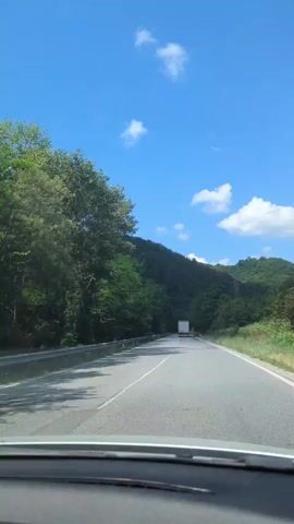 This video depicts a drive along a paved road through a lush, mountainous landscape. The road is bordered by a guardrail on the left and dense green trees and vegetation on the right. A white truck is visible driving ahead in the distance. The sky is a clear blue with scattered white clouds, suggesting a pleasant, sunny day. The time of day appears to be daytime, likely midday or early afternoon, given the brightness of the sunlight and the shadows cast. The setting is rural and natural, characterized by the abundant trees and the rolling hills or mountains in the background. There are no people visible in the frame. The overall mood conveyed is one of travel and peaceful journey through nature. No visible text is present in the video.