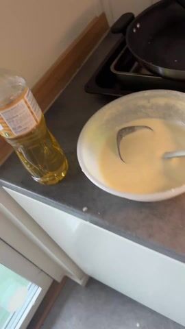 The video shows the process of making and plating crepes. 

Initially, there is a bowl of crepe batter with a ladle, and a bottle of oil on a kitchen counter next to a stovetop with a pan. A crepe is then shown cooking in the pan. The crepe is then placed on a stack of other crepes on a white plate. Next to the stack of crepes on the counter, is an open jar of chocolate hazelnut spread, with a spoon in it, and the jar's red lid next to it.