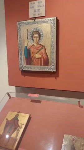 This is a video showcasing religious artwork, likely icons and relief sculptures, displayed on a reddish-orange wall in a museum or gallery. The lighting suggests an indoor setting, possibly with artificial lighting as there are no strong indicators of natural light or time of day. The scene is static, focusing on the artwork itself.

The first shot prominently features an icon of a young man with short, dark hair, wearing a red robe and a blue garment underneath, holding a spear and a palm frond. He has a halo and is framed by an ornate, gilded border. Above this icon is a plaque with Cyrillic text, which appears to be a title or inscription related to the artwork. The following shots pan across a collection of religious artworks, including a small painting of a figure on horseback battling a dragon, and several relief sculptures depicting robed figures, some with halos. Further along the wall, there are more icons, some appearing older and more stylized.

Below the wall display, glass-covered cases are visible, containing more items which are difficult to discern clearly due to the video's resolution and focus, but appear to be further artworks or artifacts laid flat. The floor is tiled with what looks like terracotta. In the background, architectural elements like brick arches and modern structural beams can be seen, suggesting a multi-level building, possibly a renovated historical site. The overall impression is one of a carefully curated exhibition of religious art. The location is identified as Peccioli, Italy, which provides context for the potential cultural and historical significance of the displayed items. No specific activities or events are taking place beyond the viewing of the art. Emotions are not discernible as the focus is on the inanimate objects.