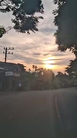 The video shows a road with a few buildings and trees on the left side. The sky is cloudy with the sun visible through the clouds. There are power lines running across the frame. A person is standing near the buildings on the left. There are also some motorbikes parked near the buildings. The camera is moving forward along the road.