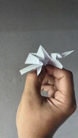 A close-up shot shows a hand holding a white paper airplane against a gray background. The airplane is folded from lined paper. The hand is positioned in the lower right portion of the frame, with the paper airplane held between the thumb and index finger. The camera moves around the airplane, showing it from different angles.