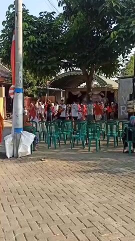 A short video captures an outdoor gathering. In the foreground, a paved walkway with brick patterns is visible. Several green plastic chairs are arranged in rows facing a raised stage area covered by a white tent. A tall, grey utility pole stands to the left, adorned with a red and white flag. A group of people, many wearing red clothing, are gathered on and around the stage, appearing to be participating in or watching an event. Some individuals, including children, are seen walking or playing on the walkway. The background shows houses and trees, indicating a residential neighborhood. The lighting suggests it is daytime, likely morning or afternoon.