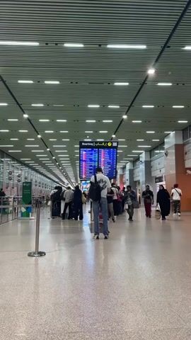 The video shows the interior of a brightly lit airport terminal. A long hallway with a high ceiling featuring parallel white panels and numerous recessed lights extends into the distance. The floor is a light-colored, possibly terrazzo, material.

Numerous travelers are present, with many people moving through the terminal or standing in lines. Several individuals are seen with luggage, including wheeled suitcases and backpacks. In the foreground, a person wearing a white t-shirt, jeans, and a black backpack stands facing away from the camera, observing a large, centrally located digital display.

This display is a flight information screen showing "Boarding" in both English and Arabic (إركاب). Below this, various flight details, such as times, destinations, and gate numbers, are visible in different colors, indicating flight statuses.

On the left side of the hallway, check-in counters are visible in the background, along with queue management stanchions in the foreground. A green sign partially reading "Economy" is also discernible on the left. People in the scene are dressed in a mix of casual attire and traditional garments, suggesting a diverse group of travelers. The overall atmosphere appears busy but orderly, characteristic of an active airport. Given the visible Arabic text and provided context, the setting is Nouaceur, Morocco.