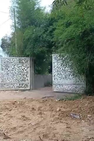 The video opens with a view of an ornate gate, partially open, leading to a dusty construction site. Beyond the gate, trees and greenery are visible. The camera then pans to the left, revealing a white scooter parked on the dirt. To the left of the scooter, a pile of black material is present, and a red plastic bag lies on the ground. Further to the left, a partially constructed building with columns and a sloped roof is visible. The environment appears to be a rural or semi-rural area with abundant vegetation, including palm trees and banana plants. The sky is overcast. The video concludes by panning further left, showing more of the construction site and the surrounding greenery.