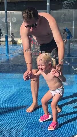 A father and his young daughter are having fun at a splash pad in Hod HaSharon, Israel. The girl, with blonde pigtails, is wearing a diaper and pink sandals. She appears to be playing with a purple wristband on her left arm. The father, shirtless and wearing patterned swim shorts, is holding onto her, likely to help her navigate the water jets. The splash pad has a blue textured surface, and water is spraying from various nozzles. In the background, other people, including children, are visible enjoying the water, suggesting a bright, sunny day. The scene is filled with a sense of joy and playful interaction between the father and daughter.