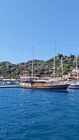 This video captures a serene coastal scene in Demre, Turkey, under a clear, bright blue sky. The foreground is dominated by calm, shimmering blue water, with gentle ripples visible on its surface.

Several boats are docked or anchored close to a lush, green hillside. The most prominent vessel is a large, traditional wooden gulet with two tall masts, positioned centrally in the frame. A Turkish flag is visible flying from its mast. To its left and right, other boats of similar style, though smaller or partially obscured, are also present, suggesting a busy harbor or tourist area.

The hillside behind the boats is densely covered with Mediterranean vegetation, including various trees and shrubs. Numerous buildings, likely houses or small establishments, are nestled into the slopes, featuring a mix of architectural styles and red-tiled roofs. The sunlight is strong, indicating a clear, warm day. The overall impression is one of tranquility and natural beauty, characteristic of a scenic coastal town. There is no discernible text visible in the video.