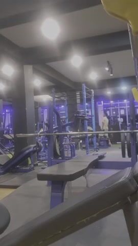Here's a detailed description of the video:

The video is set inside a gym. The camera is focused on a bench press, with a black padded bench and a blue metal frame. A barbell loaded with weights is positioned on the rack above the bench. The gym is filled with various other exercise machines, many of which are also blue.

There are a few people present in the gym. One person is walking in the background, while another seems to be using a machine further away. The gym is well-lit, with a combination of overhead lights and blue accent lighting. The overall ambiance appears modern and clean.