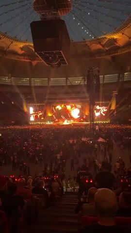 The video captures a wide shot of a large, indoor concert venue filled with a massive crowd of people. The stage, set at the far end of the stadium, is illuminated with bright, warm lights, predominantly orange and yellow, creating a fiery atmosphere. Several large screens are positioned above and around the stage, displaying visuals of the performers. Huge speaker systems hang from the ceiling, suggesting a powerful sound experience. The crowd, a sea of dark figures with occasional red lights from phones, stretches from the stage to the foreground, where a section of the audience is visible from behind, seated in tiered seating. The overall ambiance is one of excitement and anticipation for a live musical performance.