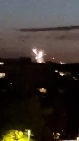 A fireworks display is captured in this short video clip. The fireworks are visible against a twilight sky, with silhouetted buildings and trees in the foreground. The fireworks ignite and explode in various patterns of light, casting a bright glow on the clouds.