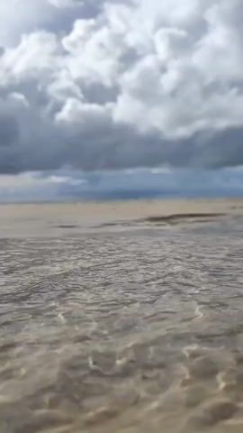 A low-angle shot of shallow water flowing over sand with ripples. The sky above is filled with dramatic, dark clouds.
