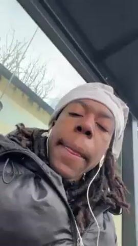A young man with dreadlocks is seated outdoors. He is wearing a white patterned headscarf, black puffer jacket, and black pants. He has white earbuds in his ears. He is holding a small green and white object in his hand. The man is sitting under a shelter, with a gray wall and windows behind him. The ground is gray with a yellow line. The camera is tilted upward and moves slightly. The man looks at the camera with a neutral expression.