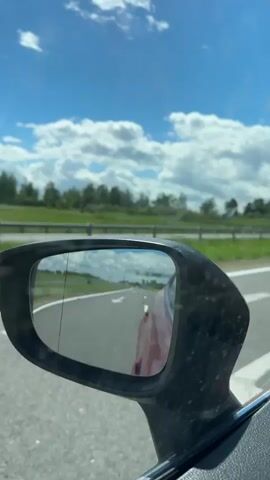 A car travels along a road. The side mirror reflects the road behind and a pink or red car. The road has white lines marking the lanes and a letter ‘E’ painted on the asphalt. On the right, there is a grassy area and a line of trees. Above the trees is a bright blue sky with fluffy white clouds.