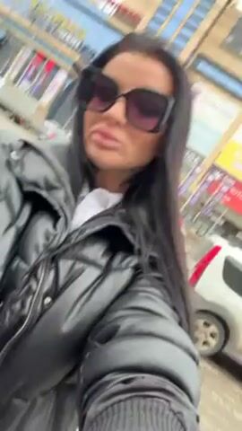 A woman films herself in a selfie-style video. She has dark hair, wears large sunglasses and a black puffy jacket over a white shirt. She appears to be walking on a sidewalk in a city, as cars and buildings are visible in the background. The sky is overcast.