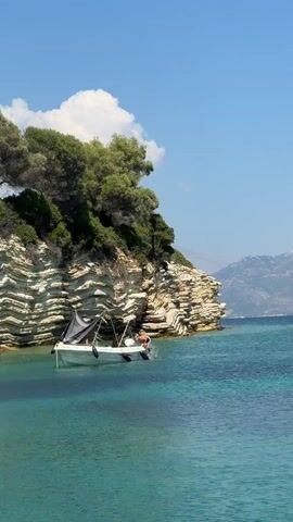 A small white boat with a dark canopy is anchored near a rocky cliff covered in greenery. Two individuals are in the water near the boat, with one appearing to be swimming or floating, and the other is partially submerged near the stern of the boat. The water is a clear turquoise color, and the sky is blue with a few white clouds. In the background, distant mountains are visible.