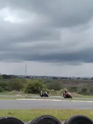 Two motorcycles are racing on a track. The first motorcycle is mostly black with red wheels and accents. The rider is wearing a black and white suit. The second motorcycle is black with a mostly red suit. The track is surrounded by grass and tires. In the background is a somewhat cloudy sky, hills, and buildings in the distance.