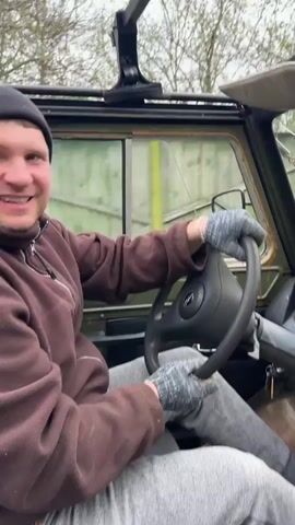 A man in a brown jacket, gray sweatpants, gloves and a beanie is driving a green off-road vehicle.  The vehicle has a black steering wheel, a black dashboard with various gauges, and a manual gearshift. The vehicle also has a rearview mirror. The windshield wipers are turned on and the windshield is cracked. The vehicle is driving down a small road with trees, houses and a red fence lining the road.