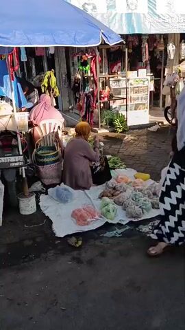 A street market scene with several people and produce. In the foreground, a woman wearing a light blue hijab and a white mask is standing and looking towards the camera. She is wearing a black and white patterned dress. To her left, a woman in a pink hijab is sitting on a white chair, facing away from the camera. In the middle ground, a woman wearing a brown hijab and a brown cardigan is sitting on the ground, sorting through green vegetables. Various produce, including carrots, potatoes, ginger, and green beans, are displayed on white cloths on the ground. In the background, there are stalls with clothing and other goods, and more people are visible walking through the market. A mosque dome is visible in the distance. The ground is paved with bricks.