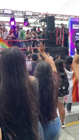 The video shows a crowded outdoor event with a stage set up. On the stage, several people are standing, some appearing to be performers or hosts. There are speakers and lighting equipment on the stage. A large rainbow flag is visible draped over the stage. In front of the stage, a large crowd of people is gathered, many of them holding up their phones to record or take pictures. Some individuals in the crowd are also waving their hands. The event appears to be a parade or festival. The lighting is bright, and the sky is visible, suggesting it's daytime.