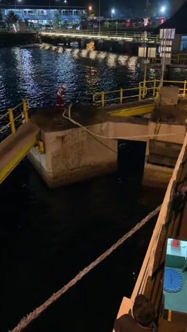 A nighttime scene at a pier. A person in a red and white uniform stands on a concrete platform near the water. Yellow ramps connect to the platform, leading away from the water's edge. In the background, a brightly lit pier with buildings and a street can be seen across the water. The foreground shows the side of a vessel, with a thick rope extending across the frame. Two individuals are visible on the vessel, one wearing a dark shirt and the other in a light-colored shirt and a cap, working with a bucket. The water is dark and reflects the lights from the distant pier.