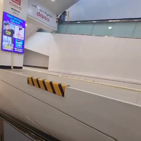 The video shows an escalator or moving walkway inside a commercial establishment. The walkway is lined with yellow and black hazard stripes along the side. A large screen displaying advertisements is visible in the upper left corner. Further up the walkway, there are signs and posters, including one for "Daiso." A person is seen at the top of the walkway. Closer to the camera, a man is pushing a yellow shopping cart with a young child sitting in it. The man is looking back and appears to be speaking. Another person is visible in the foreground, partially obscuring the view. Several other individuals are further up the moving walkway. The environment appears to be well-lit with overhead lighting.