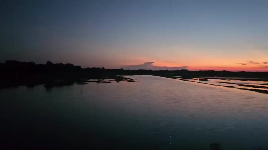 A wide shot captures a serene body of water under a twilight sky. The horizon is painted with hues of orange and red from a setting sun, transitioning to a deep blue overhead, with a single star visible. Dark silhouettes of trees line the distant banks of the water, with hints of structures or vegetation visible in the mid-ground. The water itself is calm, reflecting the sky's colors and appearing dark as night descends. The overall scene conveys a peaceful and tranquil atmosphere.